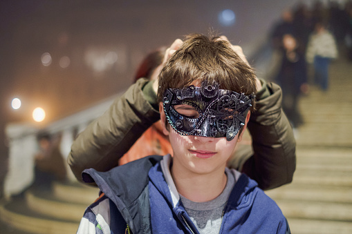 Portrait of teenage boy wearing mask, his sister is tying the string at the back of his head, family visiting Venice, Italy on winter vacation at time of Carnival