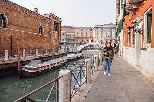 Beautiful teenage girl walking alone on stone paved pier by the canal with anchored boats, enjoying her winter break in Venice, Italy