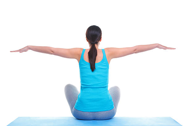 Rear view of a brunette woman sat on a yoga mat with her arms out in a balance position. Isolated on a white background.
