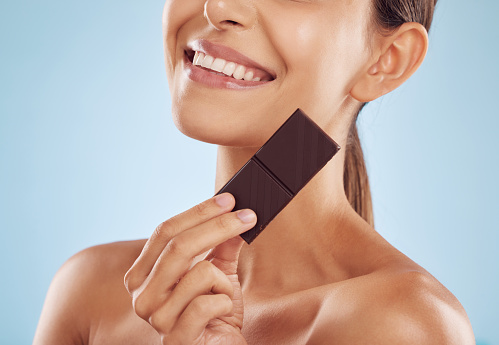 Smile, skincare and woman with chocolate for beauty isolated on a blue background. Happy, candy and model with food, cacao and dessert, sweets and sugar for eating, cholesterol and natural cosmetics