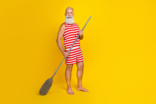 Full body size photo of professional experienced old sportsman standing on boat holding wooden oar isolated on yellow color background.