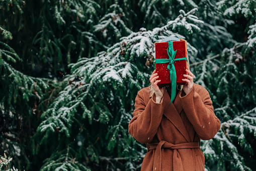 woman in brown coat hide behind with Christmas gift box in snowy winter forest, Poland