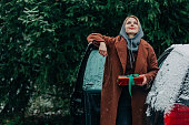 Blond hair woman in brown coat with Christmas gift box stands next to a car in snowy winter outdoor