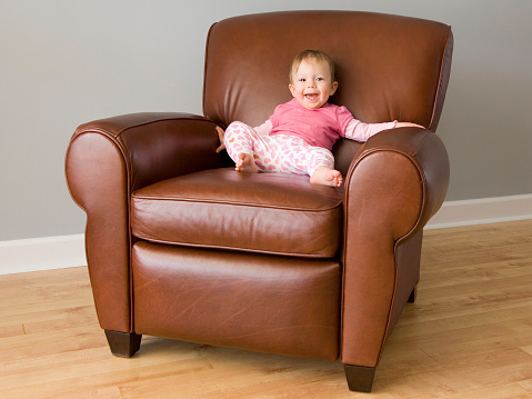 Small baby girl smiling and proud as she sits in an adult sized leather recliner chair.