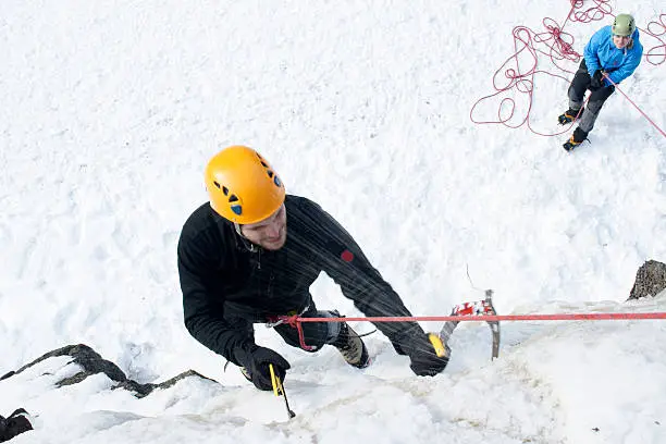 A guy climbing and a woman belaying him. The guy drives one ice axe into the frozen waterfall.