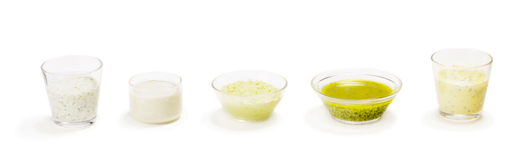 Five salad dressings, in separate glass bowls on white.