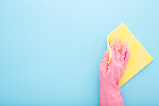Woman hand in rubber protective glove holding sponge cloth and wiping table, wall or floor surface in kitchen, bathroom or other room. Closeup. Empty place for text on blue background. Top down view.