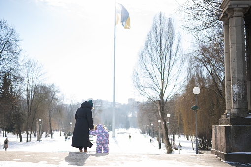 Back view of mother and child walking on a sunny frosty winter day in the park, against background of a flagpole with the Ukrainian flag.