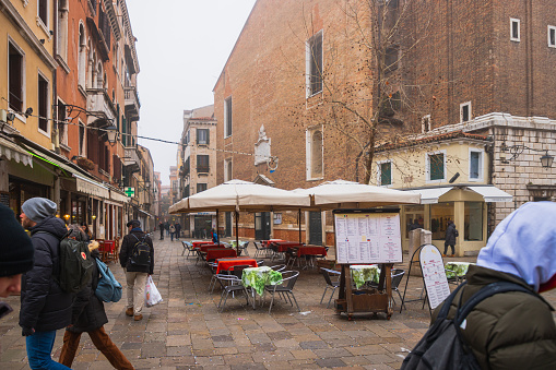 Venice, Italy - February 16 2023: Stone paved pedestrian street in Venice with sidewalk café in the middle, tourists and local people outdoors on a day in winter