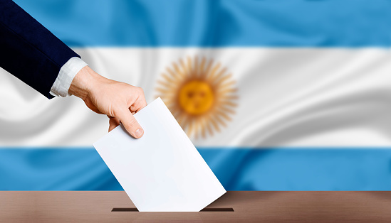Hand holding ballot in voting ballot box with Argentina flag in background. Hand man puts ballot paper in voting box on Argentina flag background. Argentina electoral elections, concept