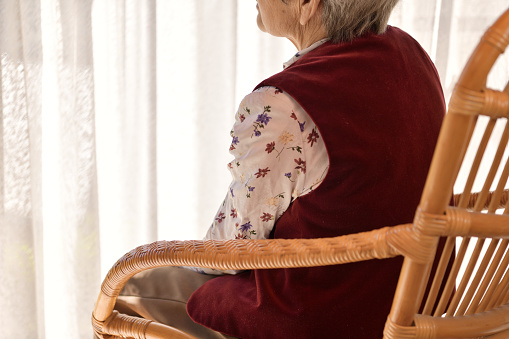 Rear view of an elderly Japanese woman wearing a red leather vest sitting on a wicker chair by the window and looking at the scenery