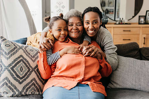 Granddaughter and mother hugging senior woman from behind and smiling cheerfully at camera on sofa at home