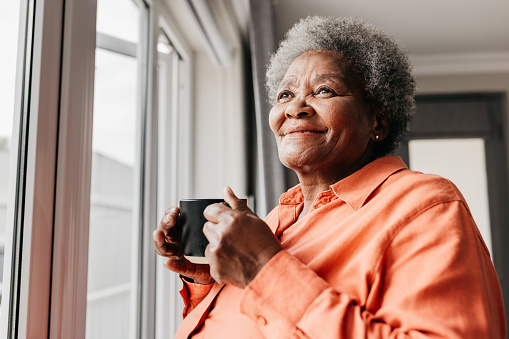 Senior woman holding mug, smiling and looking into distance beside window at home