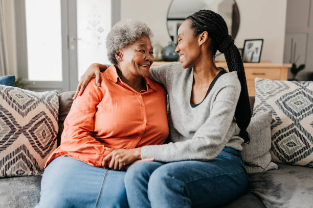 Portrait of young woman hugging happy mother on sofa at home Young woman hugging mother on sofa at home, smiling cheerfully at each other black mother and adult daughter stock pictures, royalty-free photos & images