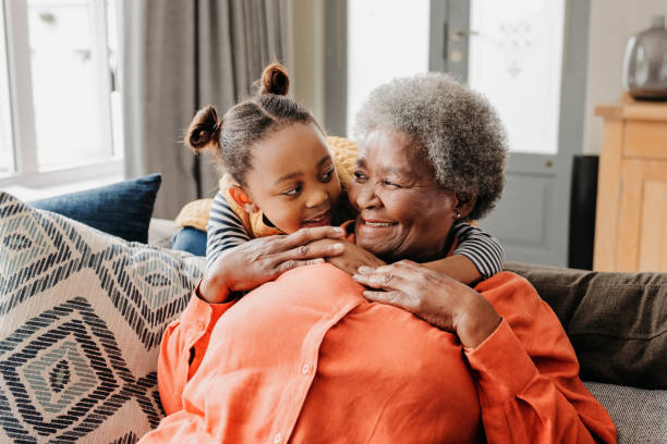 Front view of grandmother and granddaughter smiling at each other on sofa Little girl hugging grandmother from behind, smiling at each other on sofa at home pics of the black granny stock pictures, royalty-free photos & images