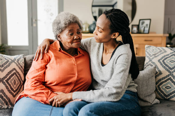 Young woman hugging and comforting mother on sofa at home Young woman hugging sad mother comforting her on sofa at home black mother and adult daughter stock pictures, royalty-free photos & images