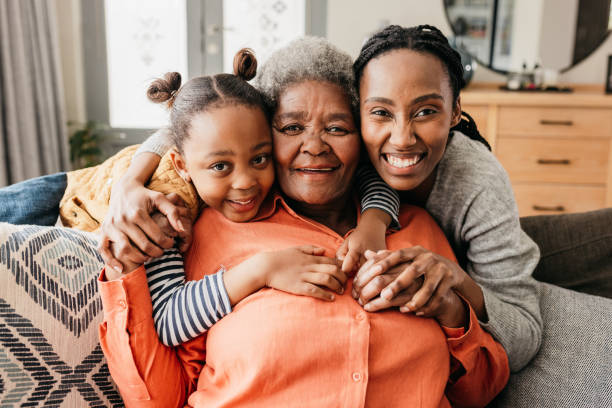 Front view of senior woman with adult offspring and granddaughter hugging and smiling cheerfully on sofa at home Cheerful granddaughter and mother hugging senior woman from behind and smiling at camera on sofa at home pics of the black granny stock pictures, royalty-free photos & images