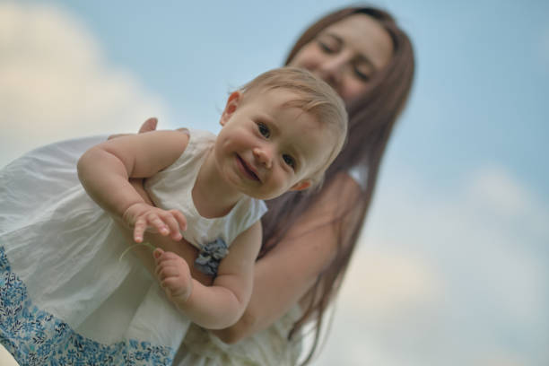 Mother lifts joyous baby in the air, under a tree canopy. Captures the joy of parenting and nature's embrace
