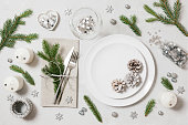 Festive table setting for Christmas dinner on white background. New Year serving with silver decor, fir branches and cones. View from above.