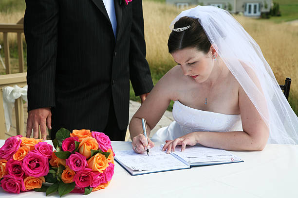Close-up shot of an unrecognizable Asian woman in a white wedding dress signing a wedding certificate with a pen on a formal document table.\n\nConcept:\nasian bride, wedding certificate signing, marriage document, bridal hands, wedding ceremony detail, close-up wedding moment, bride paperwork, wedding registry, asian wedding tradition, marriage license signing, wedding day documentation