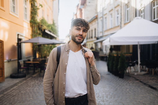 A handsome man with a warm smile stands in a charming historic European city street. His joyful expression adds a touch of charm to the picturesque surroundings.