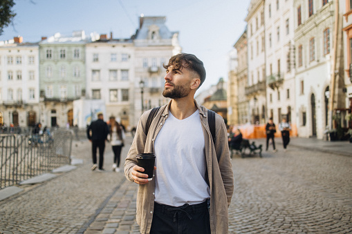 A handsome man tourist is depicted as he explores the enchanting streets of a European historical city, taking in the sights while enjoying a coffee to go, making the most of his travel experience