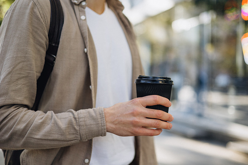 An unrecognisable man is seen carrying a coffee to go as he walks through a bustling city street, blending seamlessly into the urban scenery.