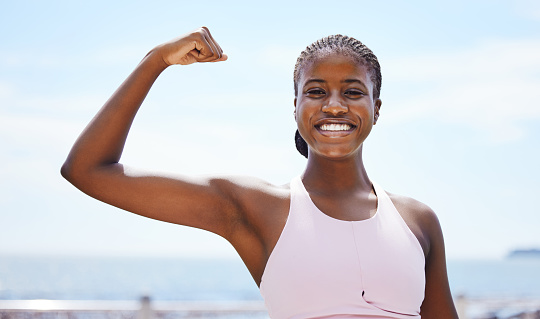 Fitness, portrait and black woman flexing muscle at the beach in summer after strength training, exercise and workout. Smile, motivation and happy girl outdoors with healthy and strong arm biceps