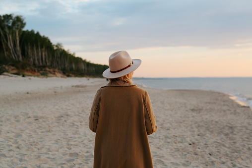 View from back of lonely senior woman in brown coat and hat walking on beach in evening, pensive adult caucasian lady alone looking at sea.