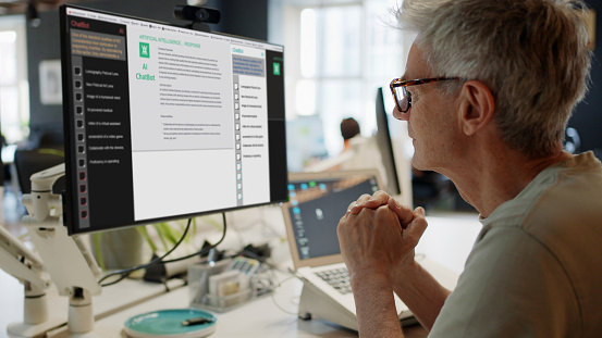 Stock image of a mature man, in an open office, looking into a computer screen as type is being added to the screen by an Artificial intelligence,  AI,  chatbot.