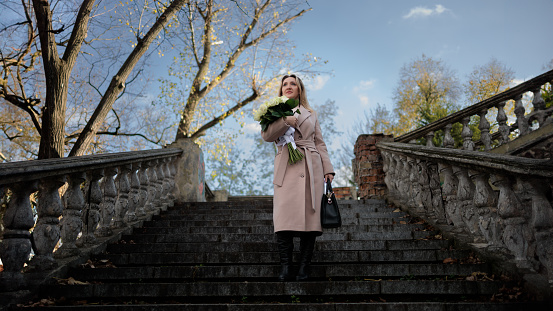 A business woman stands on an ancient staircase, holding white flowers in her hands. Stylish woman in a coat in autumn with a bouquet of white roses.