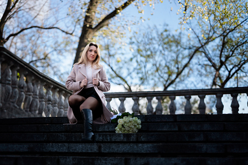 a beautiful woman sits on a stone staircase, near which stands a bouquet of white roses. Stylish woman in a coat in autumn with a bouquet of white roses.