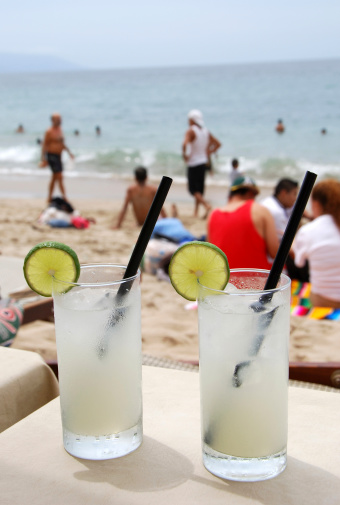 Two Margarita  cocktails at beach bar. People walking and sitting in sand.Ocean at background