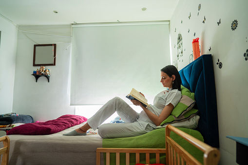 Latina woman of average age of 30 years old dressed in her medical uniform is semi-lying on her bed reading a book before going to work