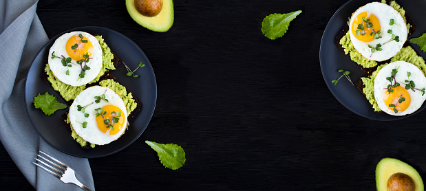 Avocado toasts for breakfast with rye bread, avocado puree, fried eggs and microgreen on the black  background. Top view. Copy space.
