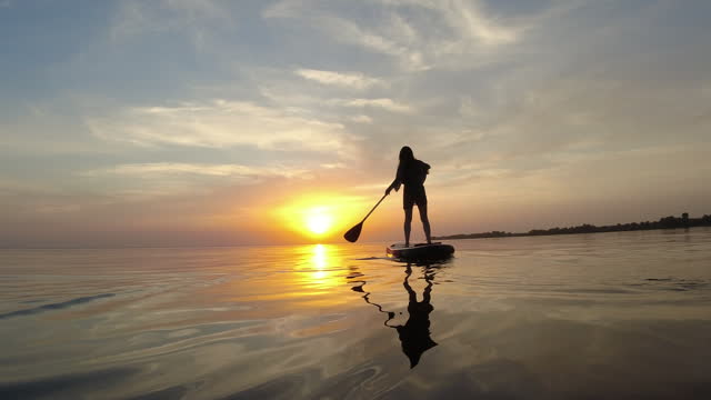 Female figure standing on a sup board and sailing into the setting summer sun. Water recreation sports