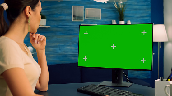 Freelancer woman at home sitting on desk table working on computer with mock up green screen chroma key display. Freelancer using isolated pc in home office for lifestyle opinions