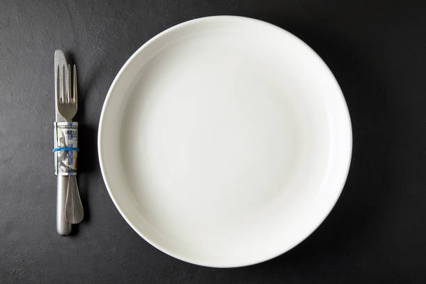istock Empty white ceramic plate with cutlery wrapped in hundred dollar bill on black stone table. Top view with copy space 1793160256