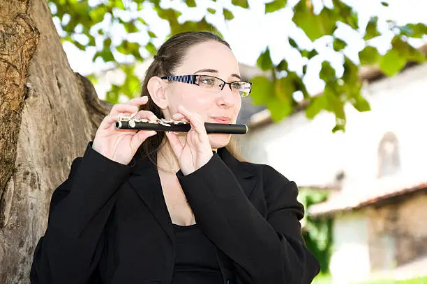 A musician with glasses, playing piccolo black with silver keys, in nature.