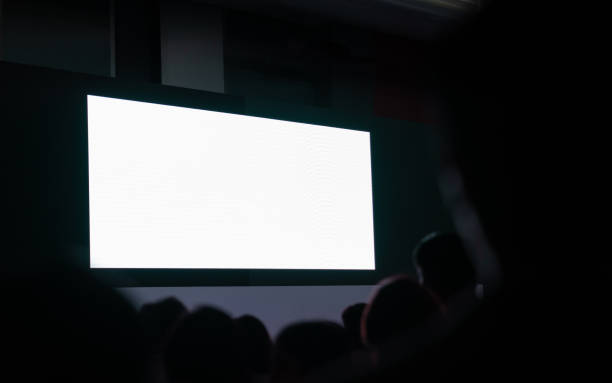 istock Front view of mockup on big blank white screen with space in empty huge hall with scene and silhouette of audiences 1793223372