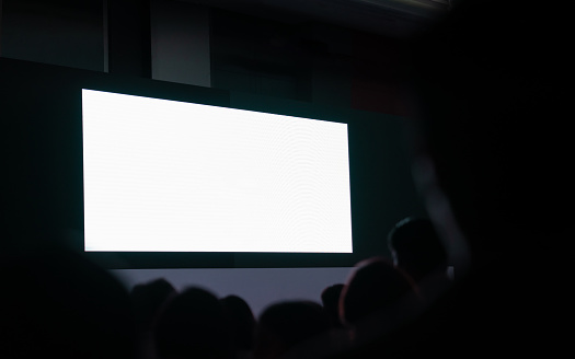 Front view of mockup on big blank white screen with space in empty huge hall with scene and silhouette of audiences