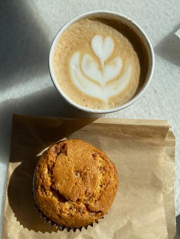 Top-down close-up view of a coffee with heart-shaped latte art in a paper cup and a sweet muffin on brown paper bag on a sunny countertop in a coffee shop in New York City