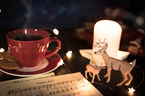 Warm toned Christmas still life with red coffee cup, carol music sheet and deer figurines in candle lit setting.