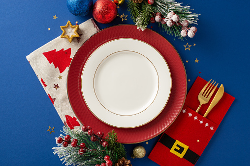Welcome New Year with touch of humor in your table setting â top view of Santa's coat-shaped holder for gold cutlery, plated, napkin, candle and festive adornments on blue background with text space