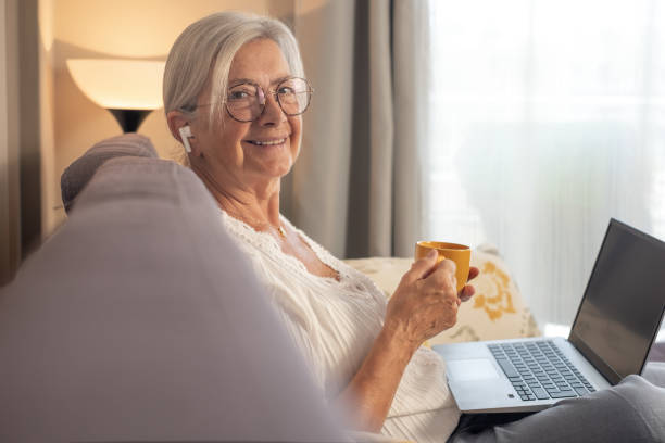 istock Relaxed handsome senior woman looking at camera sitting on sofa with a coffee cup using laptop technology listening with earphones enjoying carefree moments 1793420968