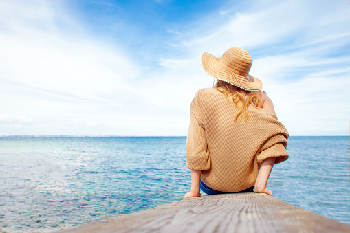 girl at the sea looks at the horizon, woman in a sun hat sits on the pier in summer and dreams, vacation concept, copy space