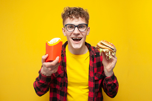 young attractive guy student in glasses holding fast food and showing surprise on yellow background, man with burger and fries
