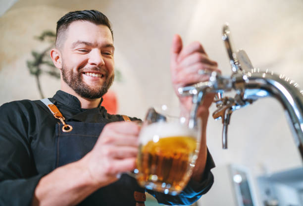istock Smiling stylish bearded barman dressed black uniform with an apron tapping fresh lager beer into pilsner glass mug at bar counter. Successful people, beer consumption, beverages industry concept image 1793447949