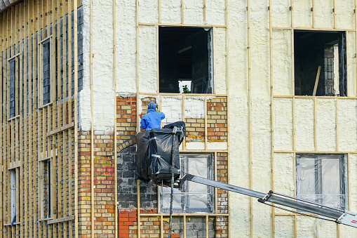 Technician spraying thermal insulation foam layer on the exterior wall of a residential house using plural component gun for polyurethane foam