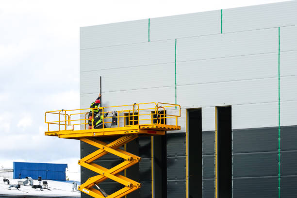 A large yellow self propelled scissor lift platform with workers against a grey sandwich panel wall Large self propelled scissor lift platform with workers at two tone grey new industrial building sandwich panels wall, industrial lifter in action sandwich panel photos stock pictures, royalty-free photos & images